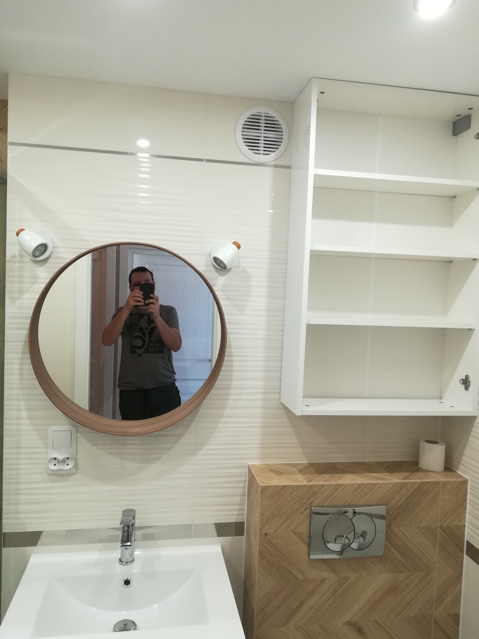 Nowoczesna łazienka z okrągłym lustrem z drewnianą ramą, białymi płytkami z delikatną fakturą, białą szafką wiszącą i umywalką z chromowaną baterią. W lustrze odbija się osoba robiąca zdjęcie.