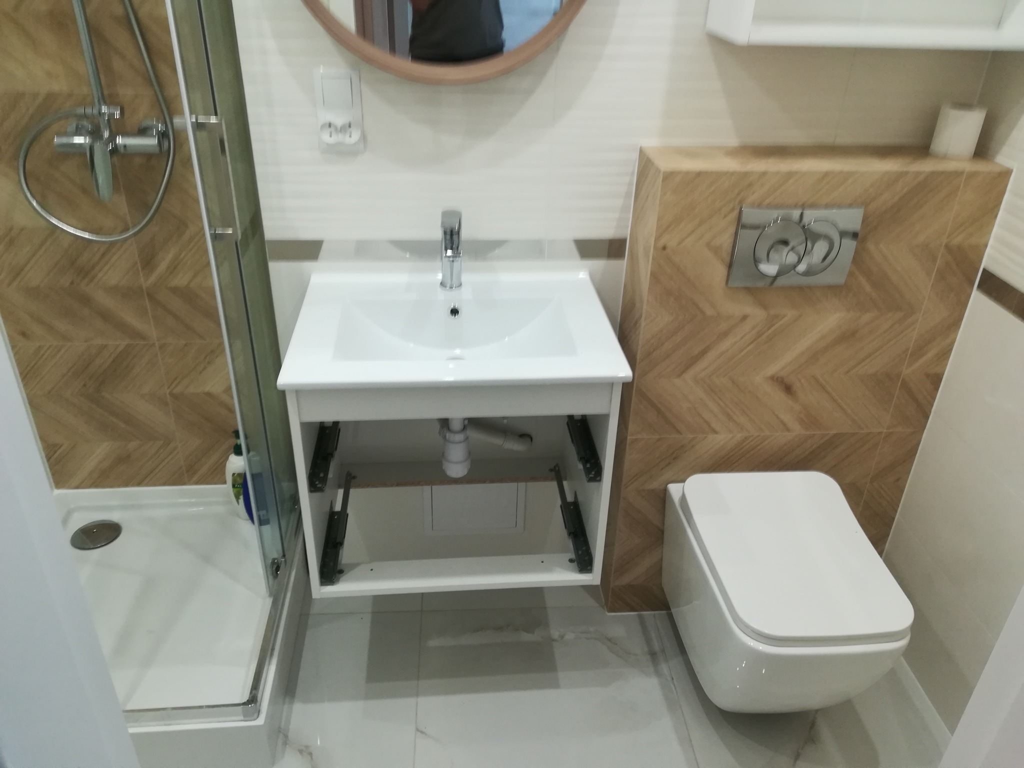 Nowoczesna łazienka z kabiną prysznicową, umywalką na szafce i podwieszanym WC, wykończona płytkami imitującymi drewno w jodełkę.