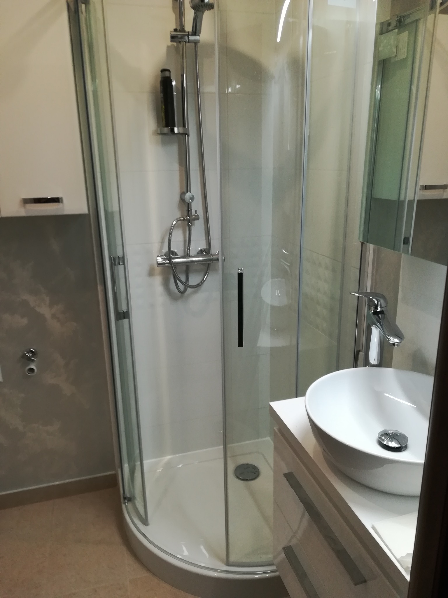 Nowoczesna kabina prysznicowa z okrągłą podstawą i szklanymi drzwiami, chromowana bateria umywalkowa nad białą umywalką nablatową, z elementami szarej ściany w tle.