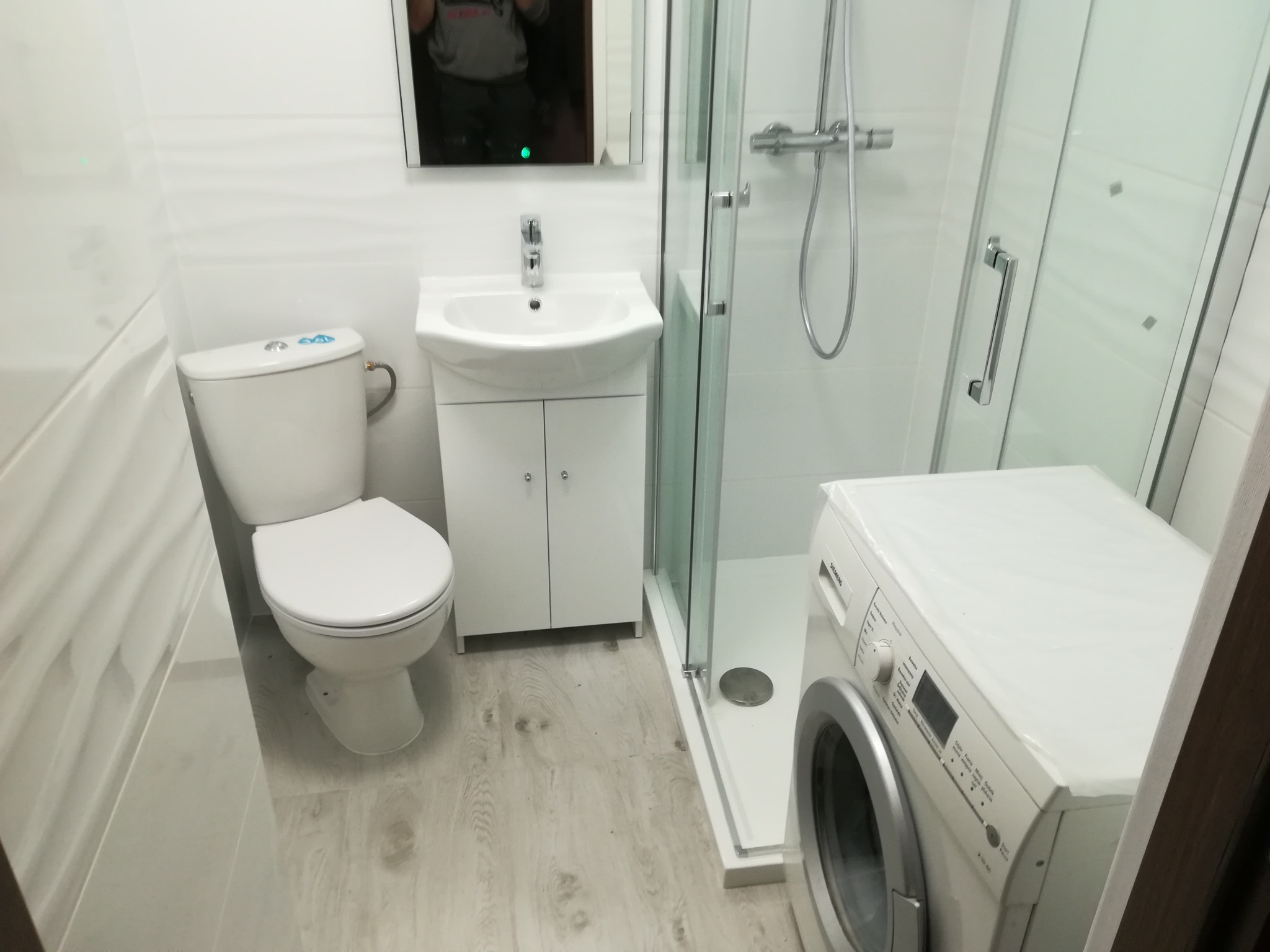 Nowoczesna łazienka po remoncie: białe płytki z delikatnym wzorem, podwieszana toaleta, umywalka z szafką, kabina prysznicowa z chromowaną armaturą i pralka w rogu, całość utrzymana w jasnej...