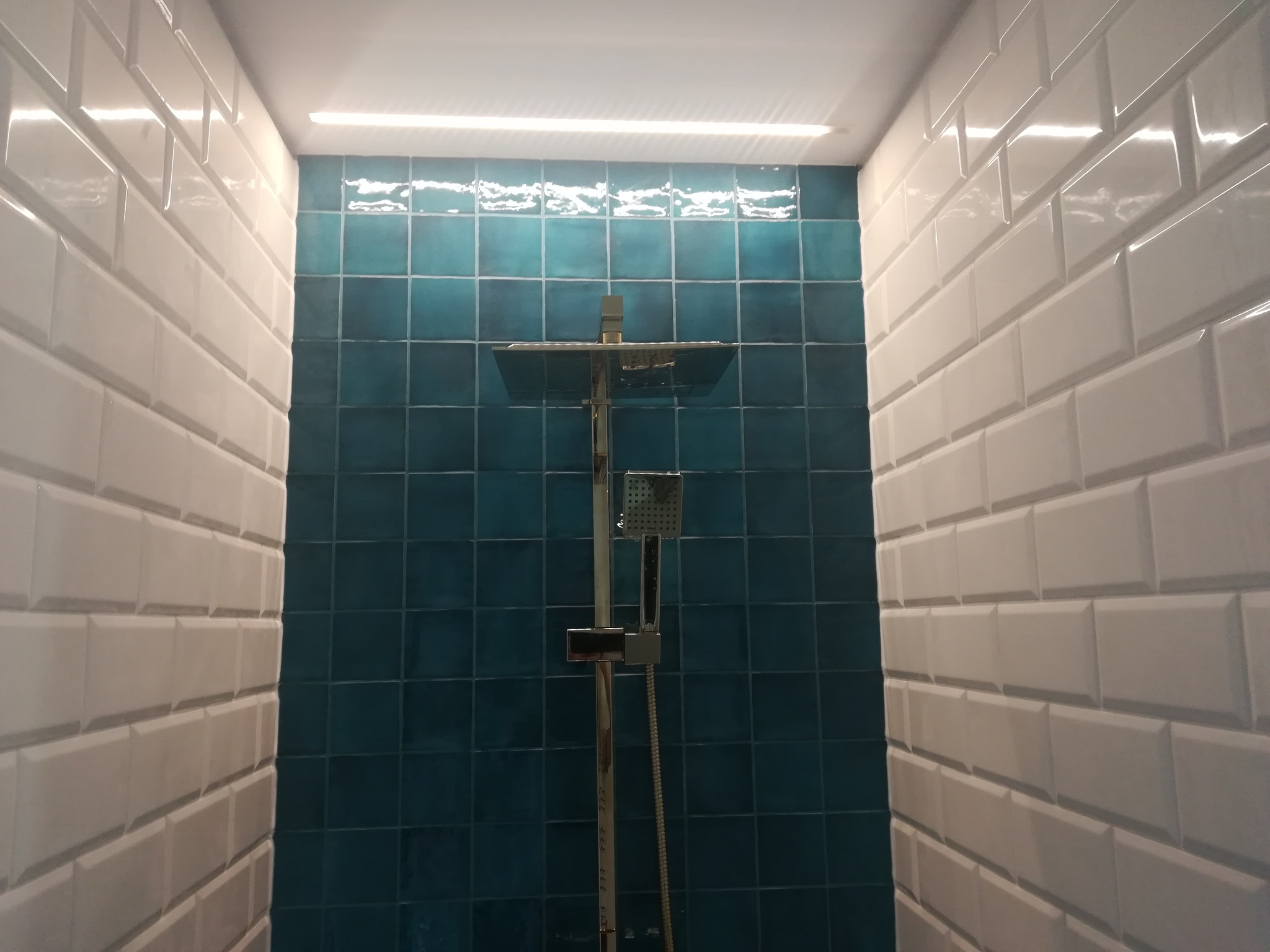 Kabina prysznicowa wykończona białymi, prostokątnymi płytkami typu metro na ścianach bocznych i turkusowymi, kwadratowymi płytkami na ścianie tylnej, z zamontowaną chromowaną armaturą natryskową.