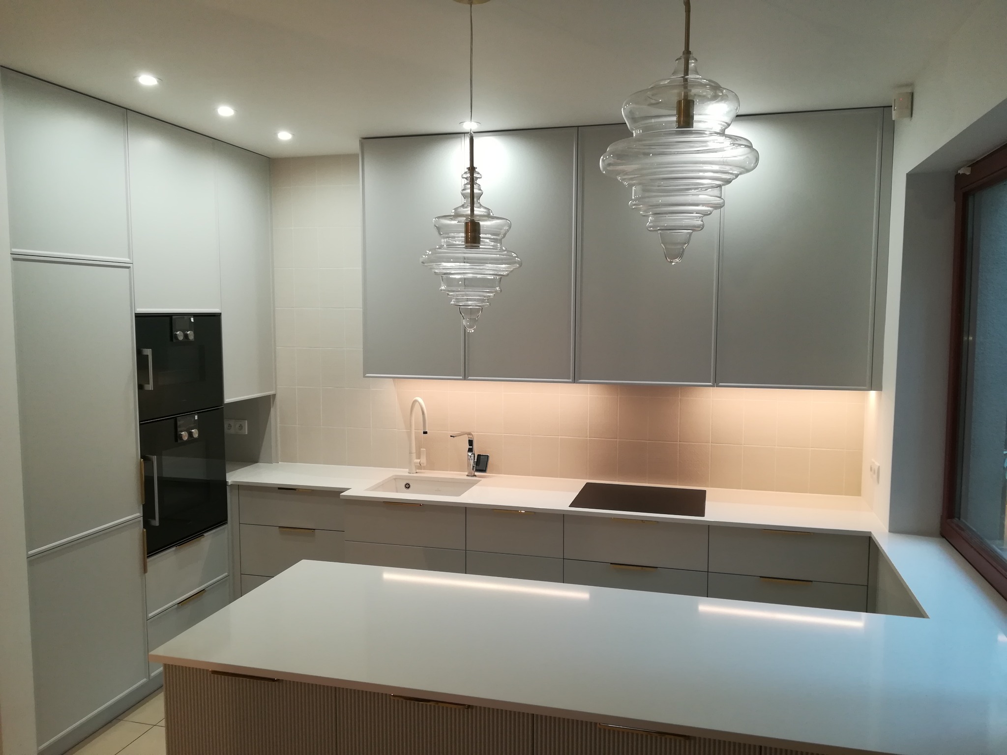 Nowoczesna, jasna kuchnia z wyspą, oświetleniem podszafkowym i dekoracyjnymi, szklanymi lampami wiszącymi.
