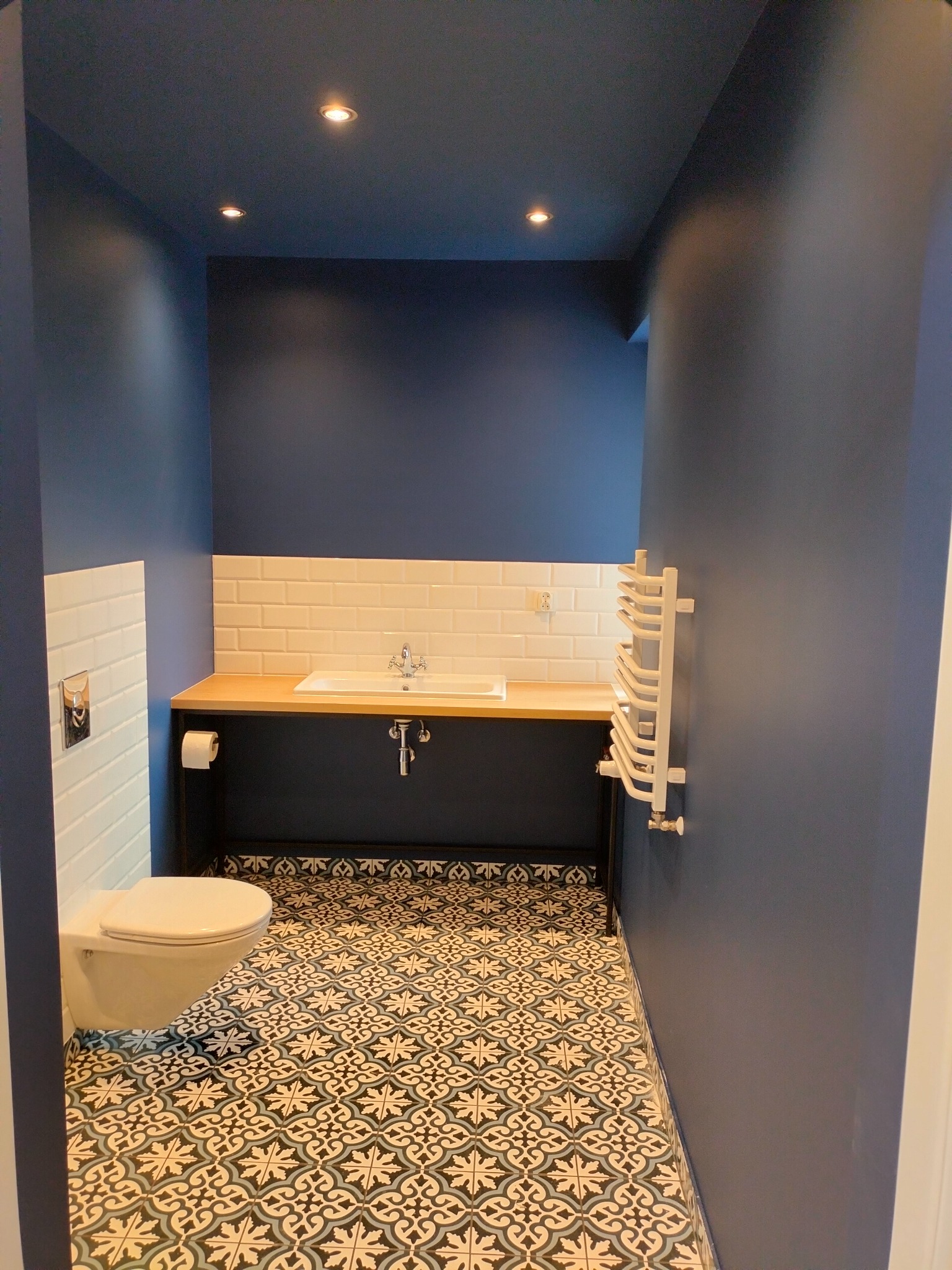 Niebieska łazienka z wzorzystą podłogą, białą armaturą i drewnianym blatem pod umywalką, oświetlona punktowymi lampami sufitowymi.