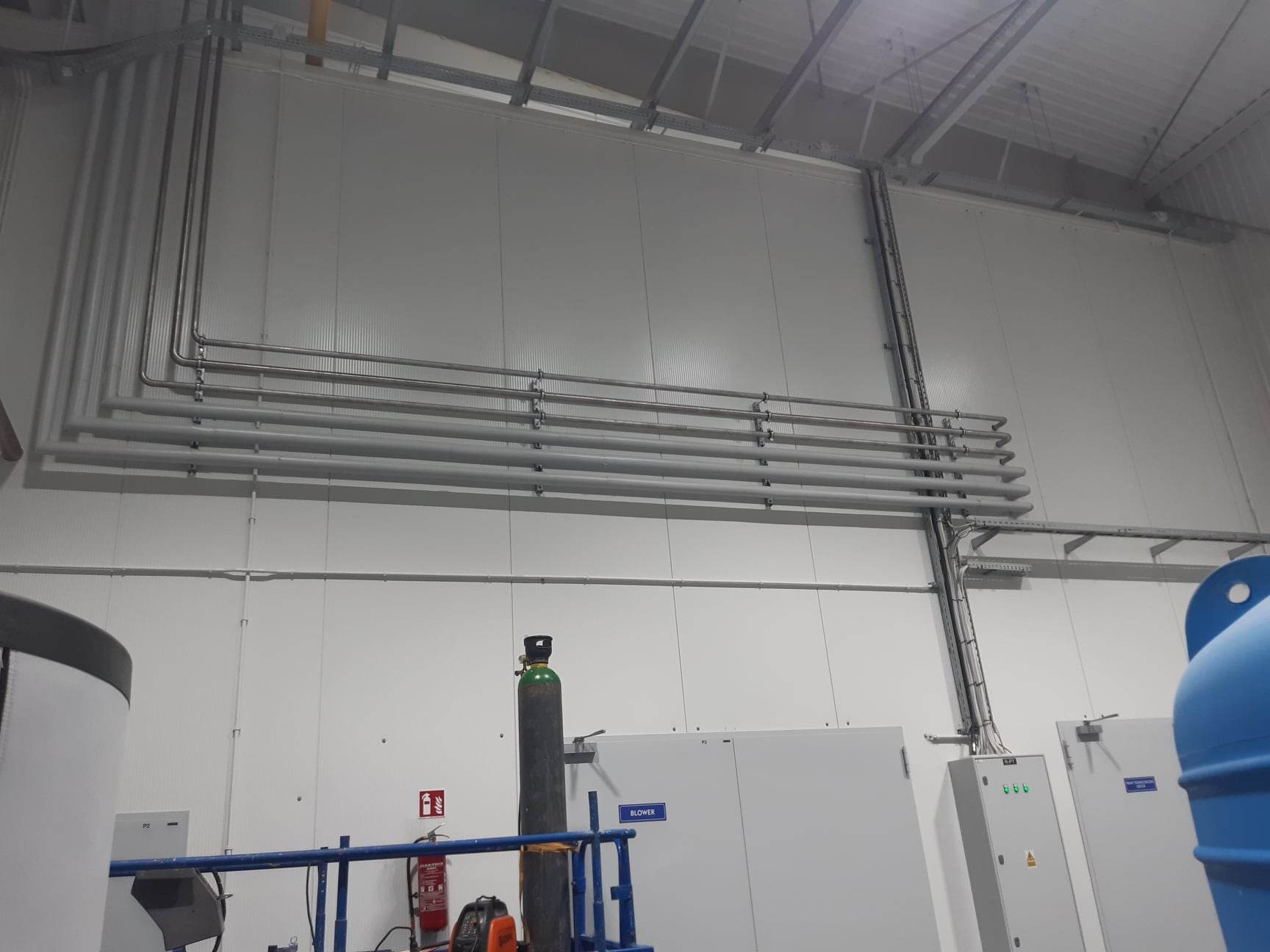 Instalacja rurociągów ze stali nierdzewnej w hali produkcyjnej, widoczny zbiornik, butla z gazem i szafa sterownicza.