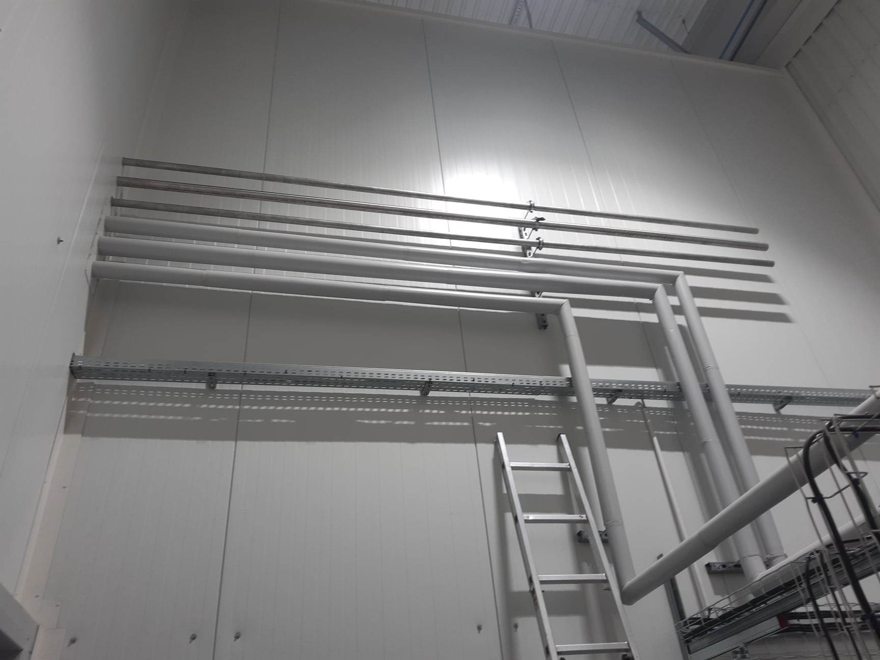 Wnętrze hali przemysłowej z instalacją rur i koryt kablowych zamontowanych na ścianie z paneli, widoczna drabina aluminiowa.