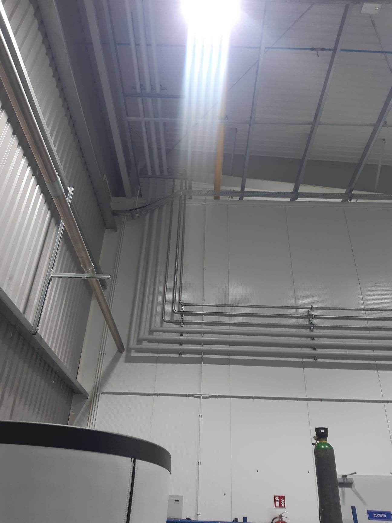 Wnętrze hali produkcyjnej z widocznymi rurami instalacji przemysłowej biegnącymi wzdłuż ściany, metalową konstrukcją pod sufitem i butlą z gazem w rogu.