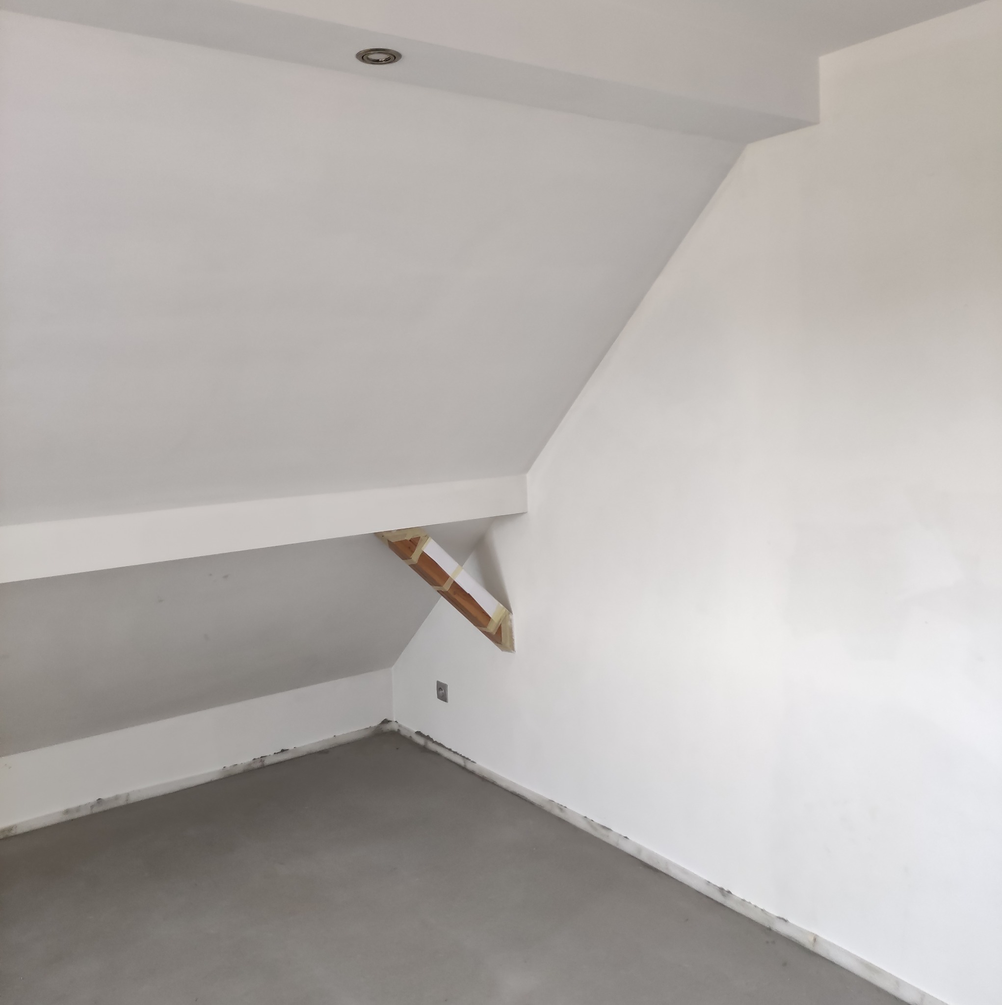 Pokój na poddaszu w trakcie wykończenia: ściany pomalowane na biało, surowa podłoga, widoczna konstrukcja drewniana z taśmą malarską i gniazdko elektryczne.