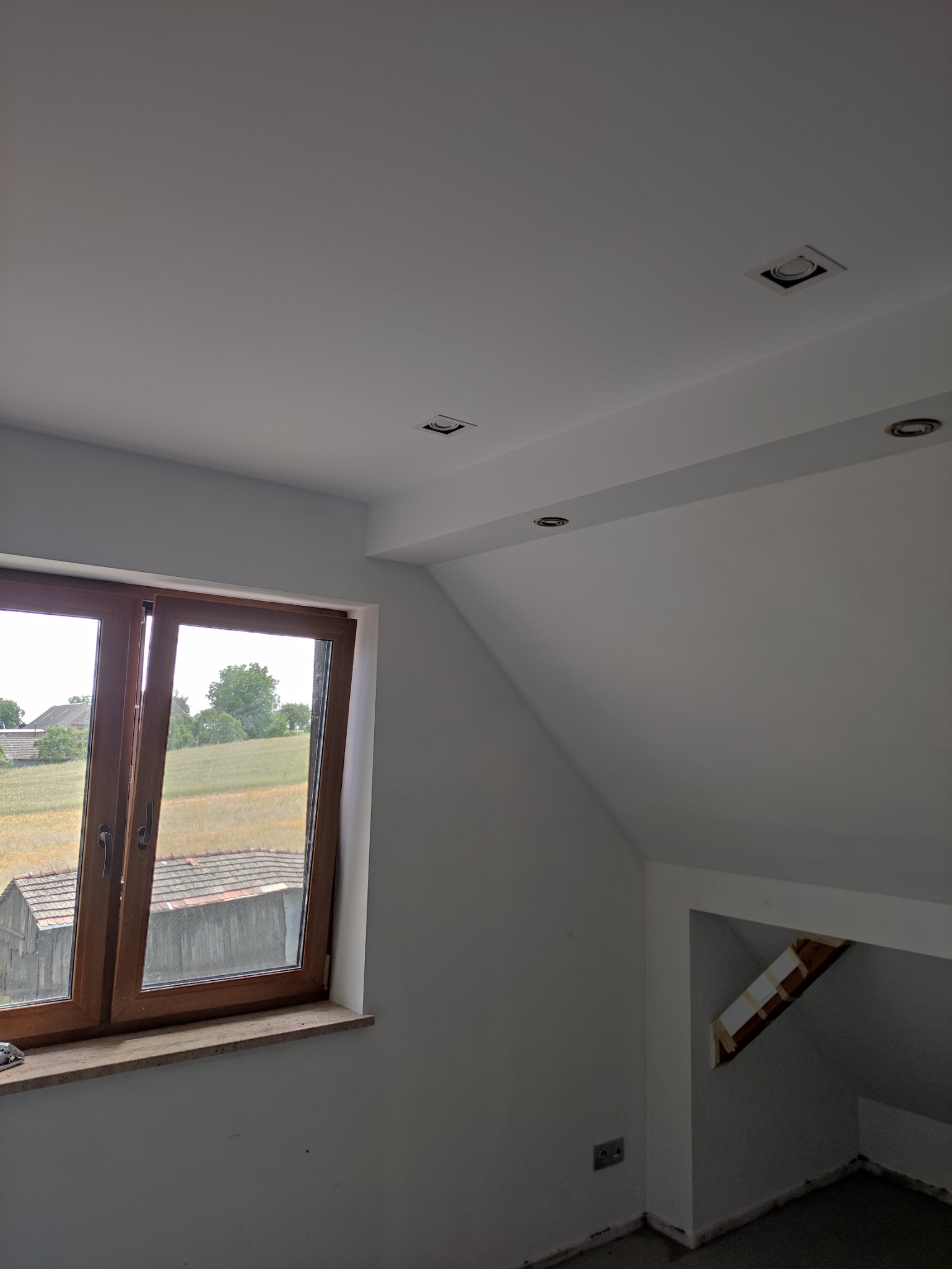 Poddasze po remoncie: drewniane okno z widokiem na wieś, białe ściany, zamontowane oświetlenie punktowe w suficie.
