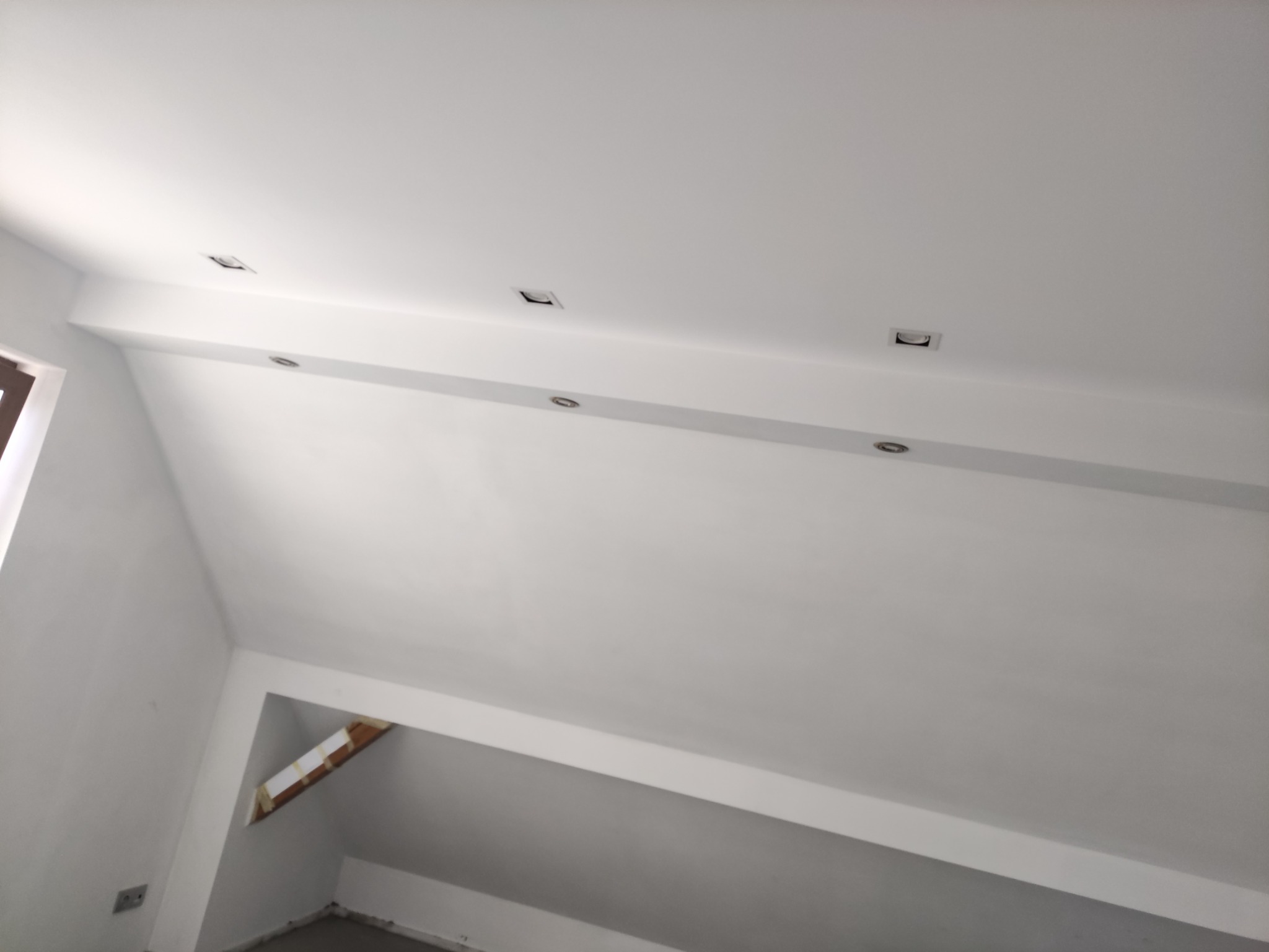Poddasze po remoncie, białe ściany i sufit ze skośnym dachem, w suficie zamontowane kwadratowe i okrągłe lampy halogenowe, widoczne fragmenty okna po lewej stronie i otworu w ścianie...