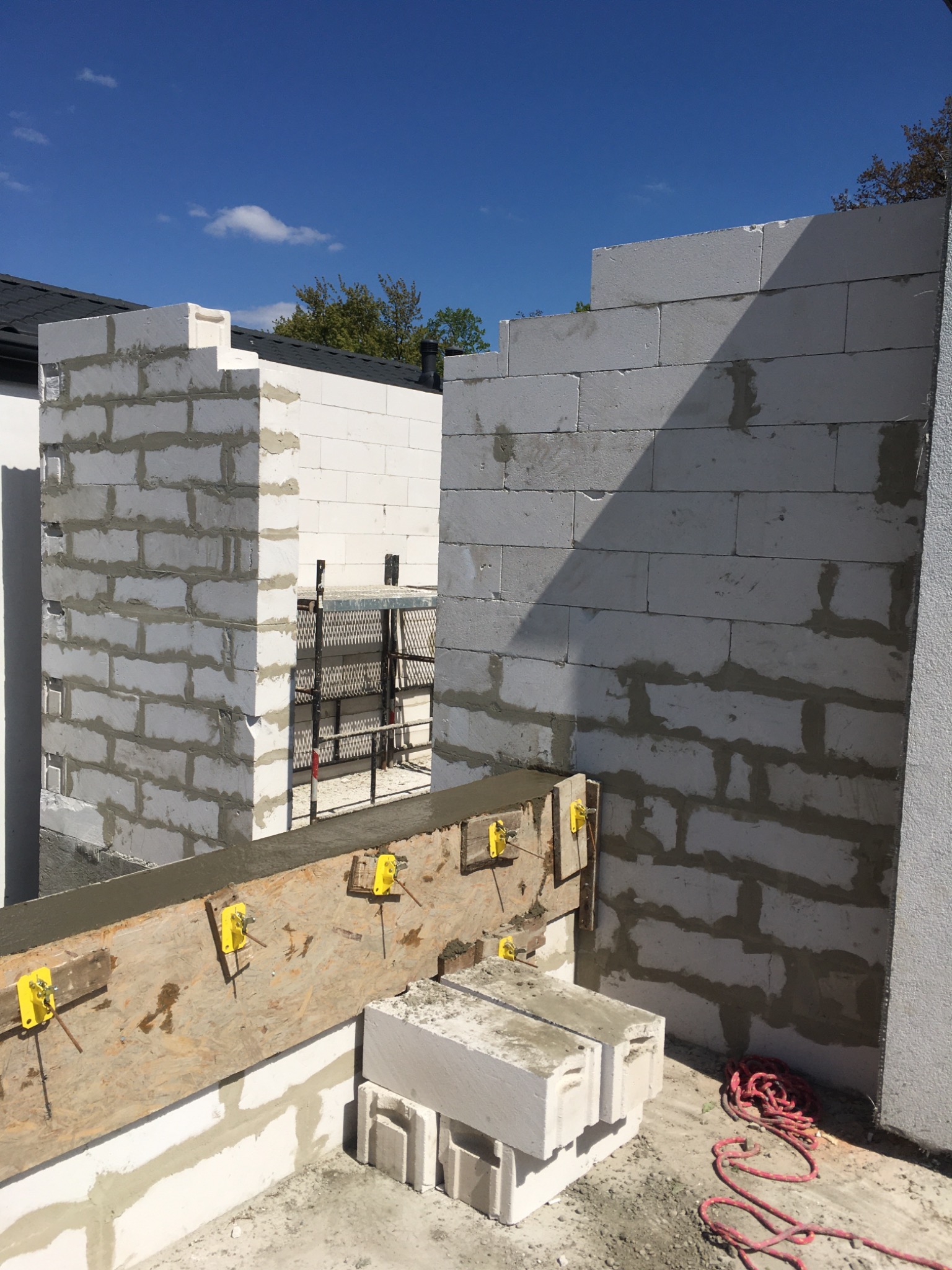 Wznoszenie ścian z bloczków betonowych, widoczne szalunki z żółtymi zaciskami, czerwona lina na podłożu, fragment dachu i błękitne niebo w tle.