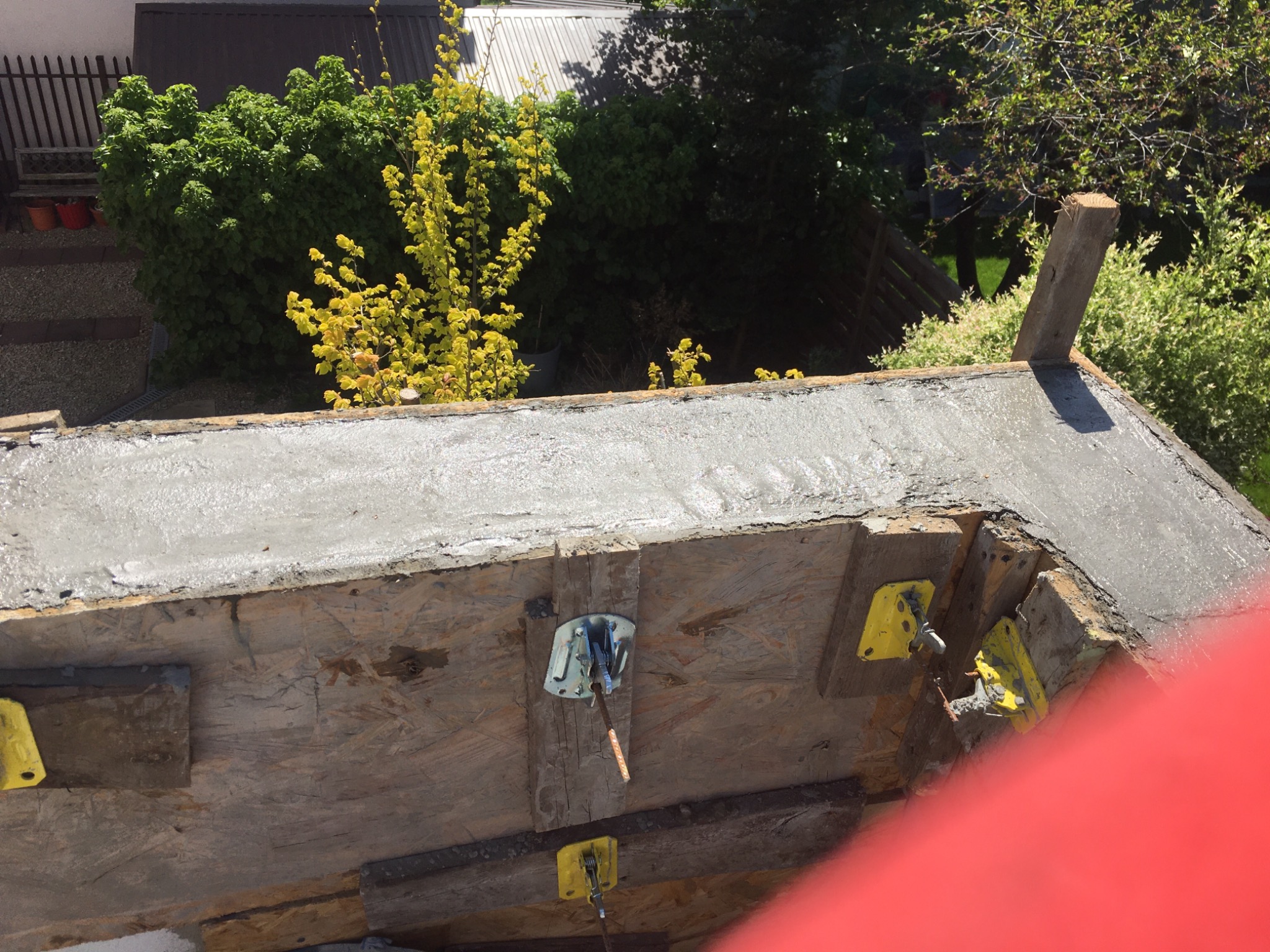 Świeżo wylany beton w szalunku drewnianym tworzącym obrys fundamentu, widoczne żółte elementy mocujące szalunek z zewnątrz, w tle zieleń drzew i fragment dachu budynku.