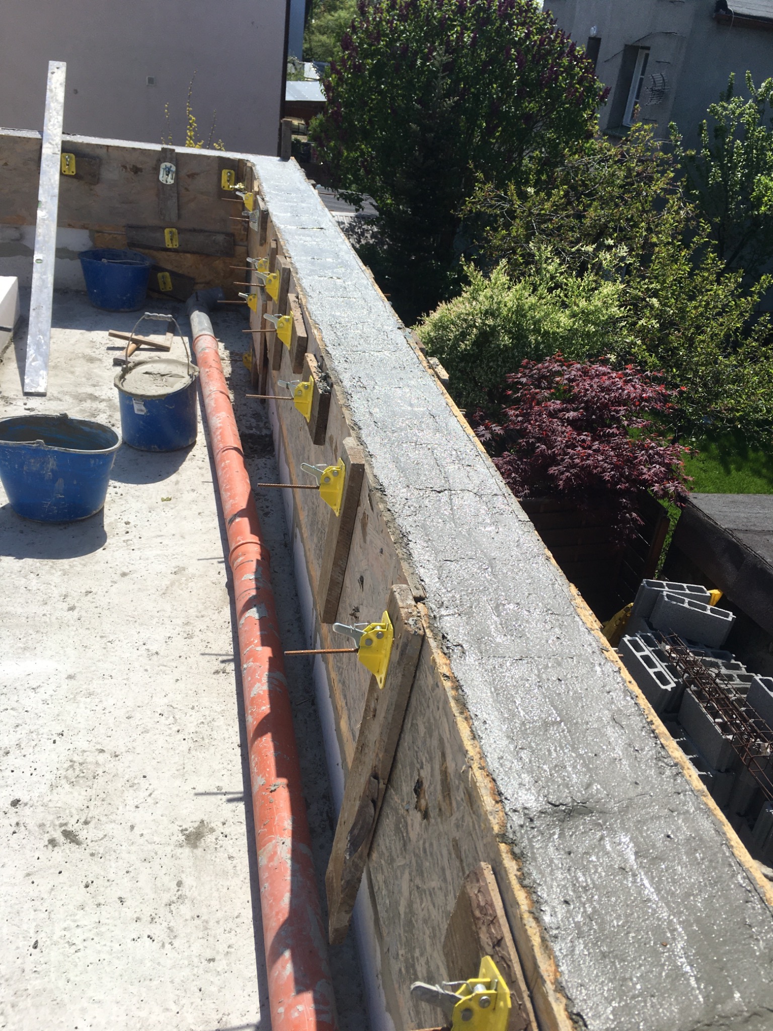 Wylany beton w szalunku fundamentowym, widoczne zbrojenie, poziomica, wiaderka i rura kanalizacyjna, w tle budynek i zieleń.