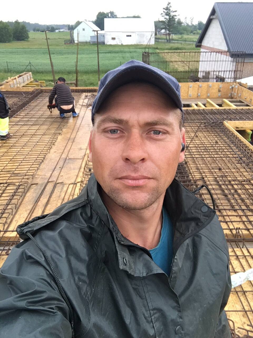 Mężczyzna w niebieskiej czapce i kurtce przeciwdeszczowej robi selfie na tle zbrojenia i szalunków, w tle pracownik przy zbrojeniu i zabudowa.
