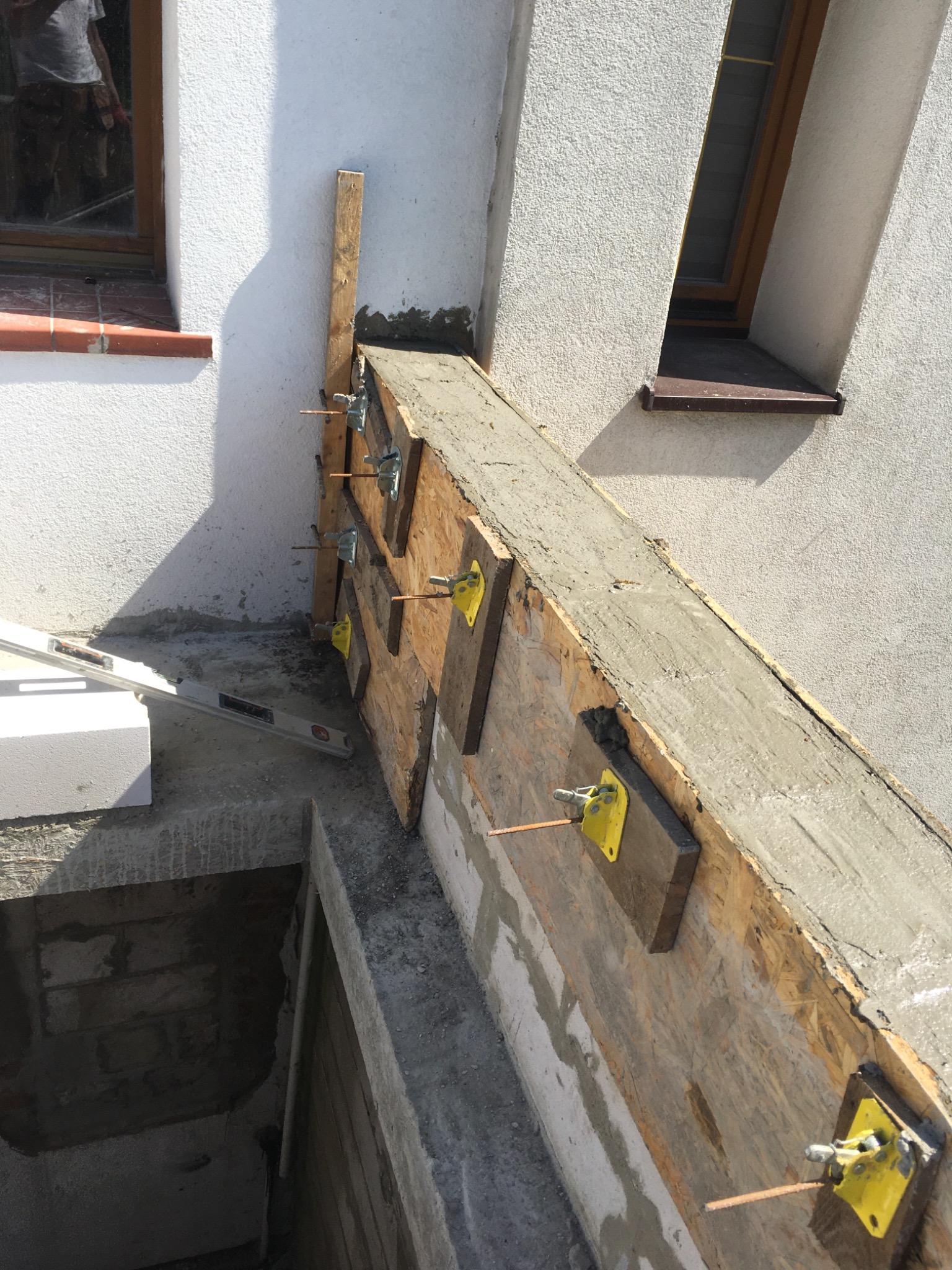 Szczegółowe ujęcie szalunku z betonu na fundamencie budynku, z widocznymi żółtymi zaciskami i wystającymi prętami zbrojeniowymi, poziomica leży obok bloczków betonowych.