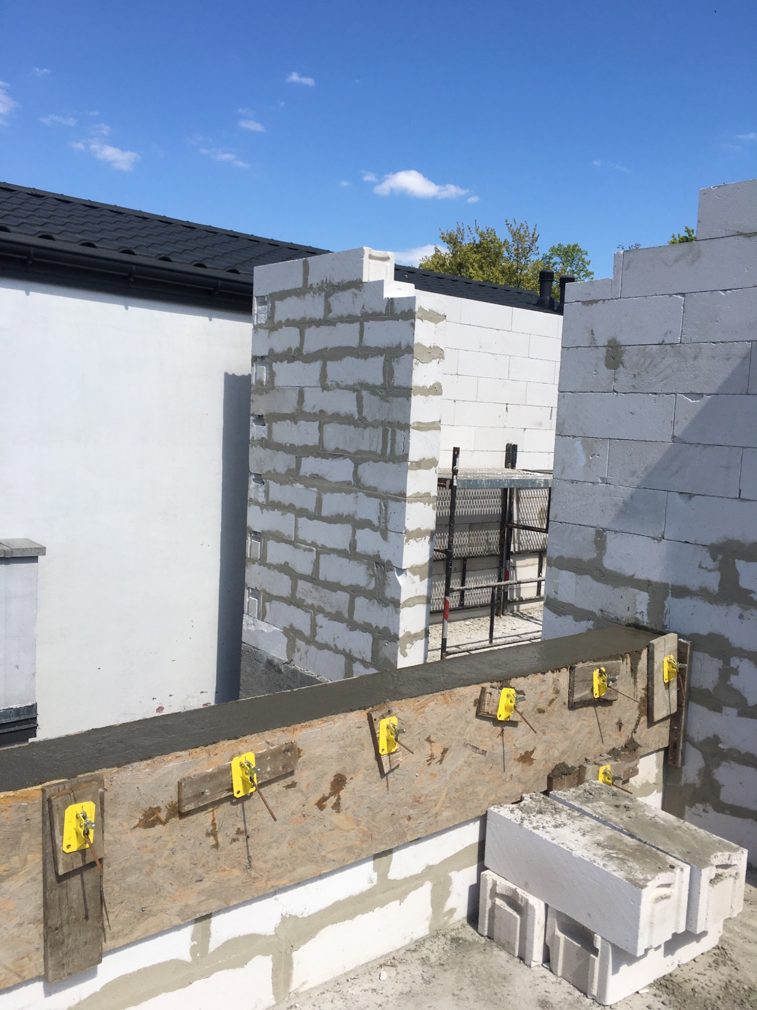 Świeżo wymurowane ściany z białych bloczków, widoczne żółte elementy mocujące szalunek, w tle rusztowanie i fragment dachu budynku.