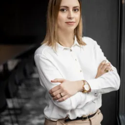 Portret kobiety w białej koszuli i beżowych spodniach, z założonymi rękami, złoty zegarek na nadgarstku, czarny pierścionek na palcu, stojącej na tle ciemnej ściany i okna.