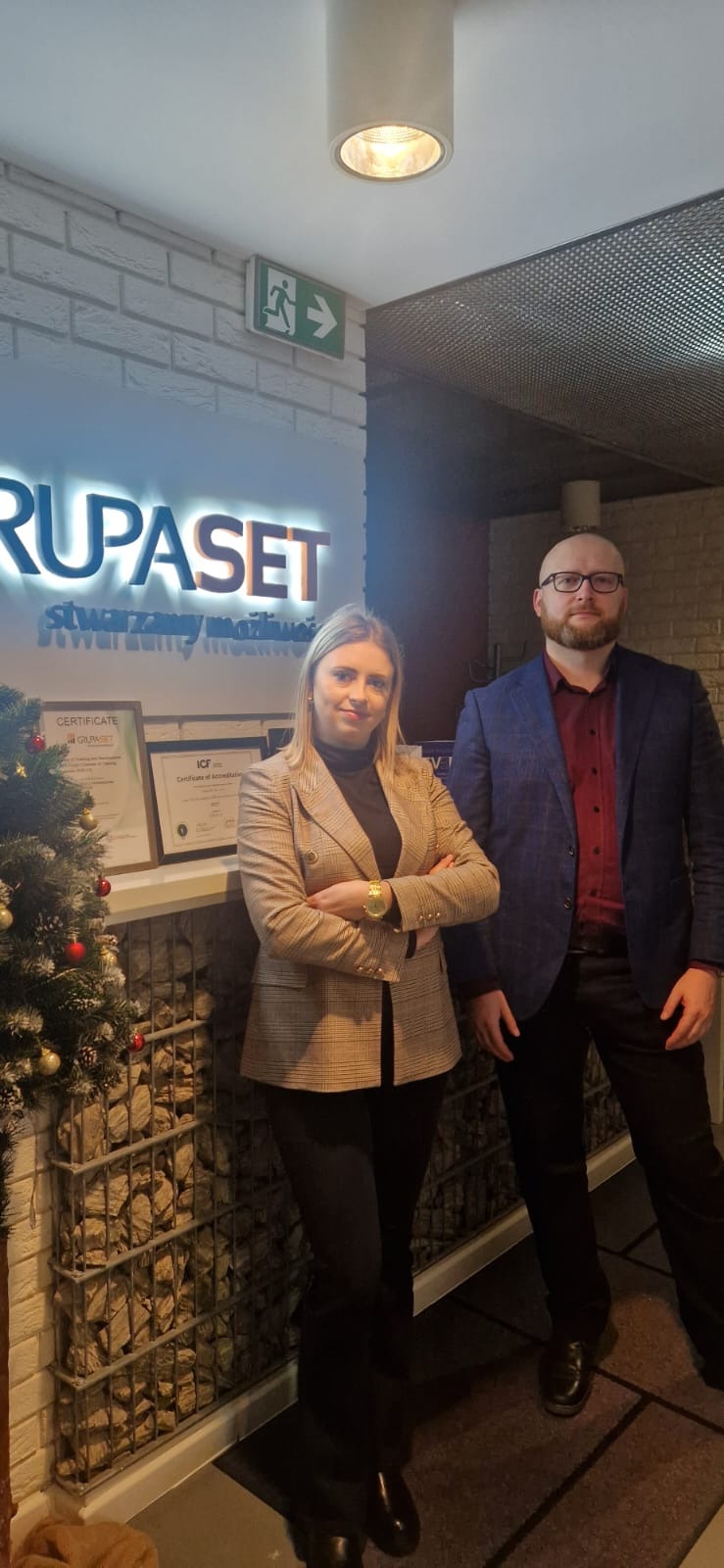 Dwoje pracowników firmy GrupaSET pozuje na tle logo i certyfikatów. Kobieta w beżowej marynarce, mężczyzna w niebieskiej marynarce. Świąteczny akcent w postaci choinki.