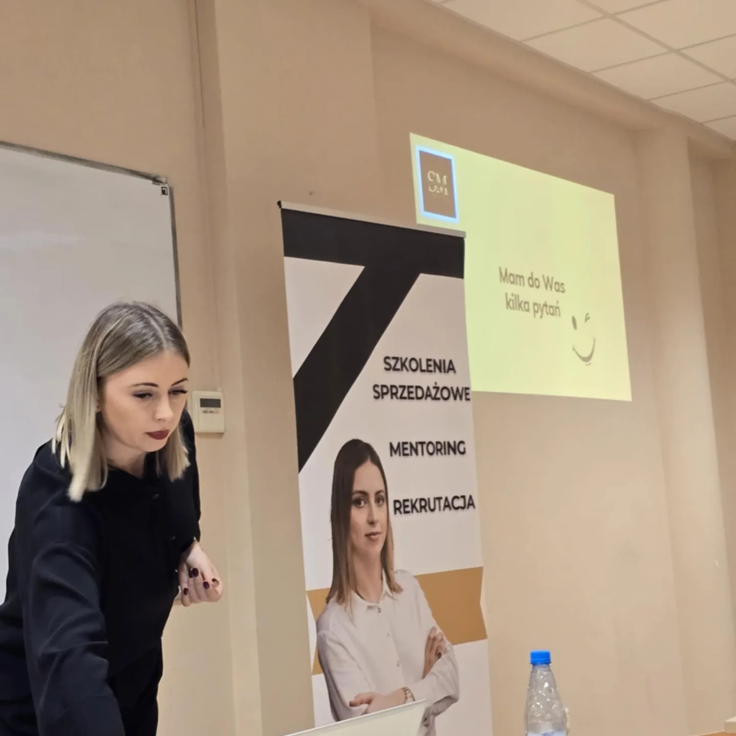 Prelegentka podczas szkolenia sprzedażowego, obok baner z ofertą mentoringu i rekrutacji. Na ścianie wyświetlany slajd z pytaniem do uczestników.