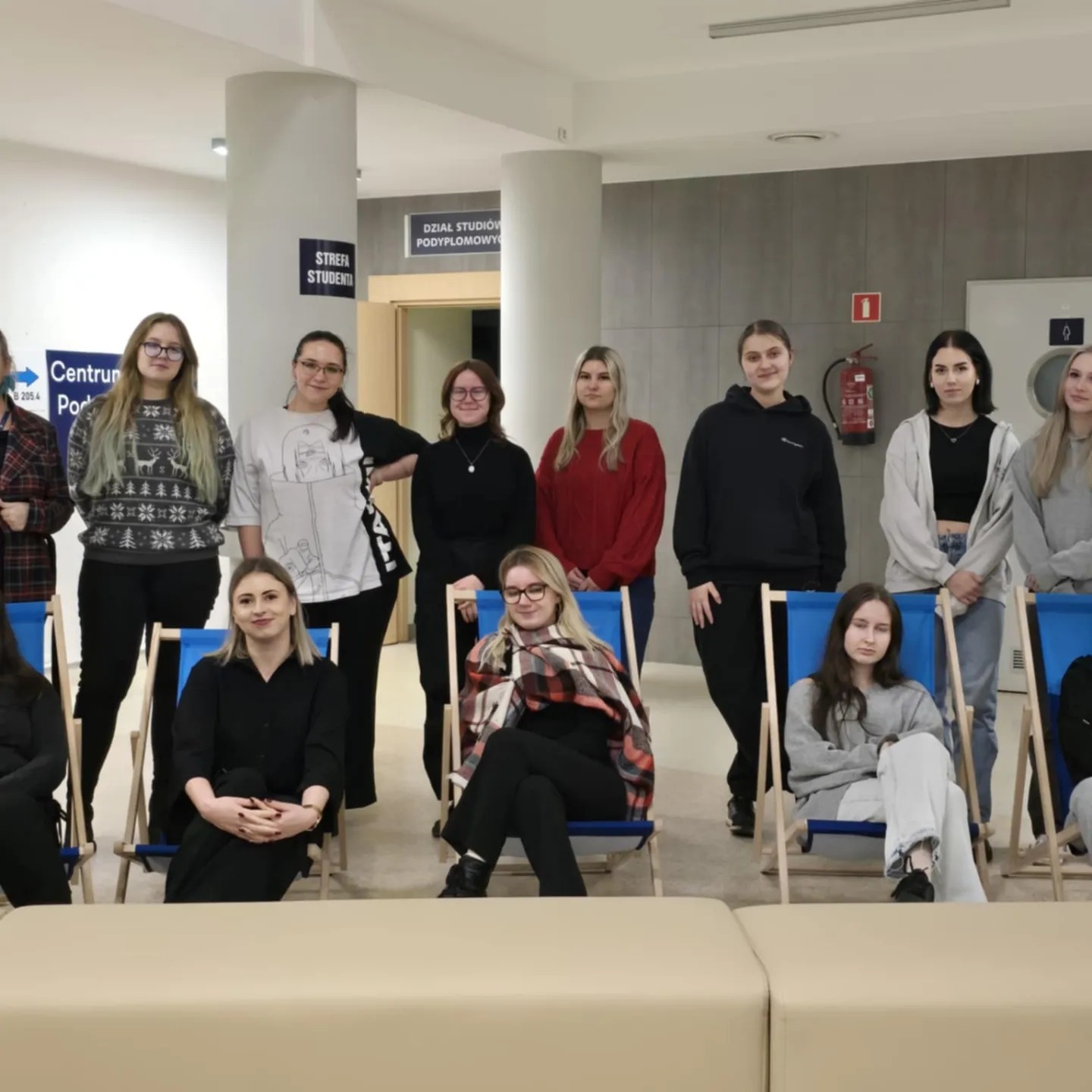 Grupa młodych kobiet, część siedzi na niebieskich leżakach, część stoi za nimi, w jasnym wnętrzu z oznaczeniami strefy studenta i działu studiów podyplomowych.