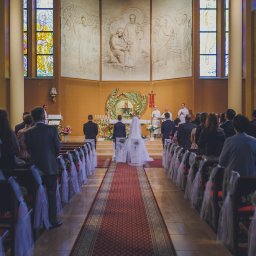 RAFIDEX Sp. z o.o. - Para młoda stoi przed ołtarzem w kościele podczas ceremonii ślubnej, widok z tyłu, czerwony dywan, dekoracje z tiulu na ławkach, goście obserwują.