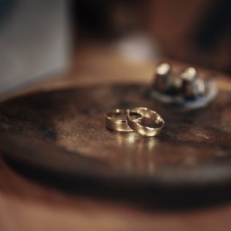 RAFIDEX Sp. z o.o. - Dwa złote obrączki ślubne leżące na okrągłej, ciemnej, metalowej tacy. W tle rozmyte przedmioty.