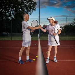 RAFIDEX Sp. z o.o. - Dwaj tenisiści, kobieta w czapce i mężczyzna, podają sobie ręce nad siatką na korcie tenisowym, trzymając rakiety, po zakończonym meczu.