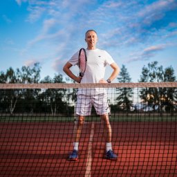 RAFIDEX Sp. z o.o. - Mężczyzna w sportowym stroju, z rakietą w ręku, stoi na korcie tenisowym za siatką, z rękami na biodrach, patrząc w obiektyw, na tle błękitnego nieba i drzew.