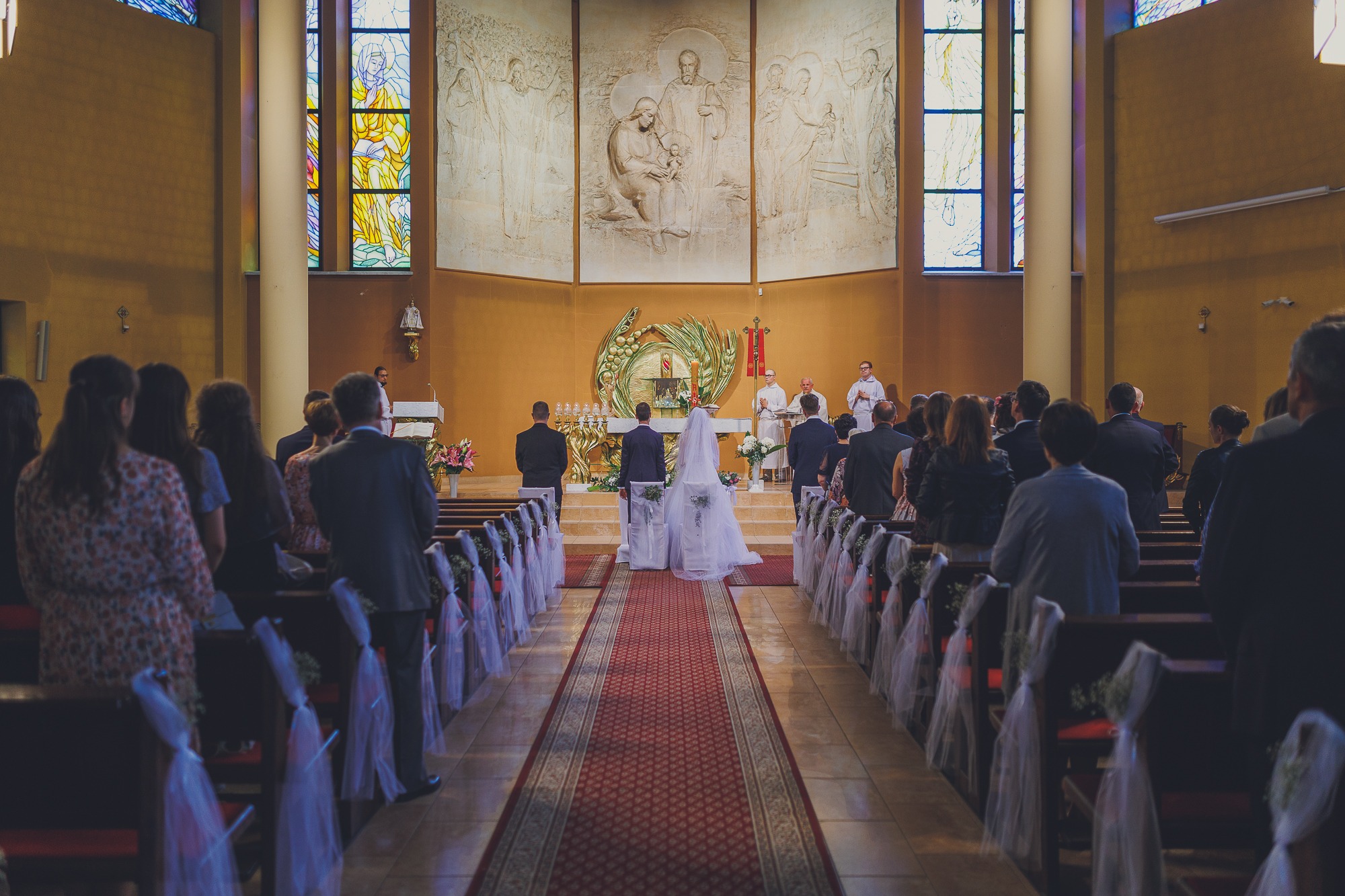 Para młoda stoi przed ołtarzem w kościele podczas ceremonii ślubnej, widok z tyłu, czerwony dywan, dekoracje z tiulu na ławkach, goście obserwują.