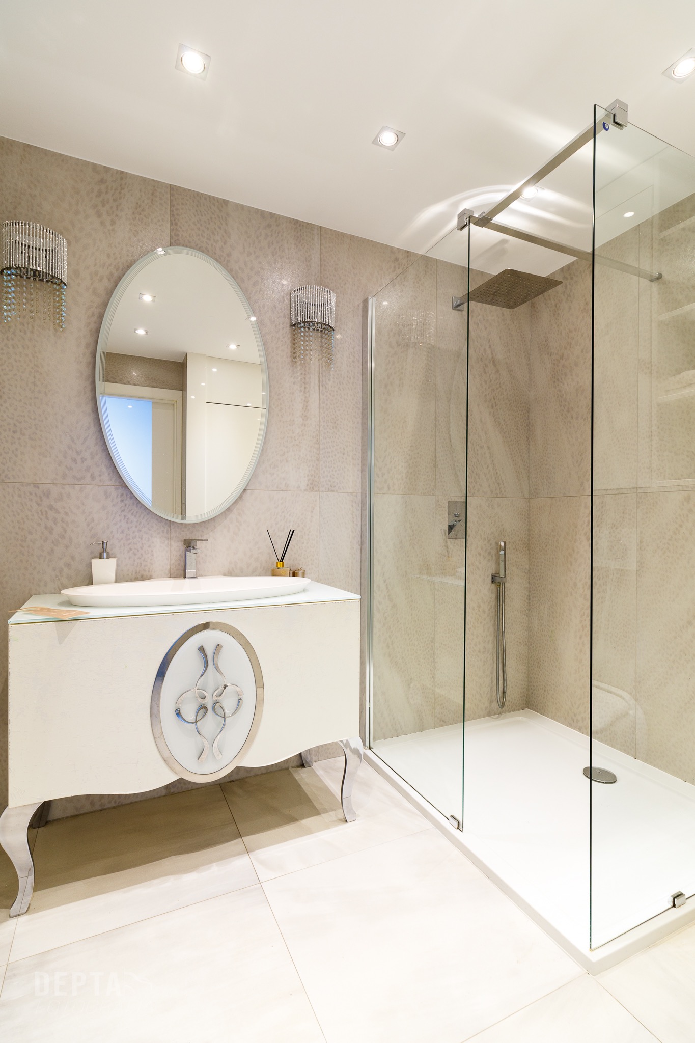 Elegancka łazienka w beżach z owalnym lustrem, ozdobnymi kinkietami z kryształkami, kabiną prysznicową typu walk-in i białą szafką umywalkową na giętych nóżkach.