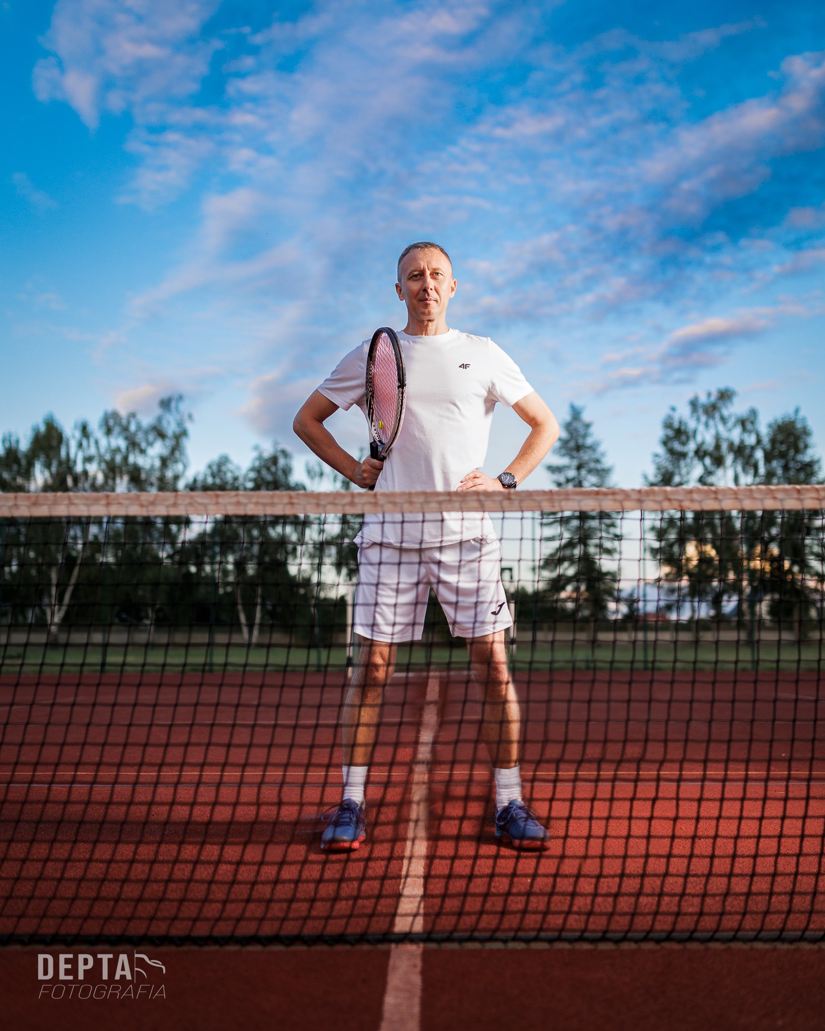 Mężczyzna w sportowym stroju, z rakietą w ręku, stoi na korcie tenisowym za siatką, z rękami na biodrach, patrząc w obiektyw, na tle błękitnego nieba i drzew.