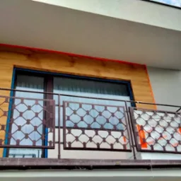 Elewacja domu z balkonem w trakcie prac wykończeniowych, widoczna drewniana okładzina, zabezpieczona folią część balustrady i pomarańczowe taśmy ochronne.