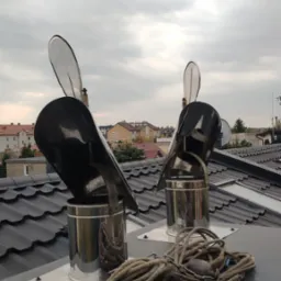 Dwa kominy z nasadami typu strażak na dachu pokrytym czarną dachówką, widoczne narzędzia pracy i zabudowa miejska w tle pod pochmurnym niebem.