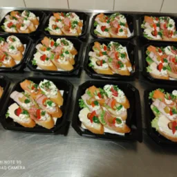 Przygotowane kanapeczki na pieczywie, udekorowane kremem, wędlinami, serem, warzywami, ułożone w czarnych, plastikowych tackach na metalowym stole.