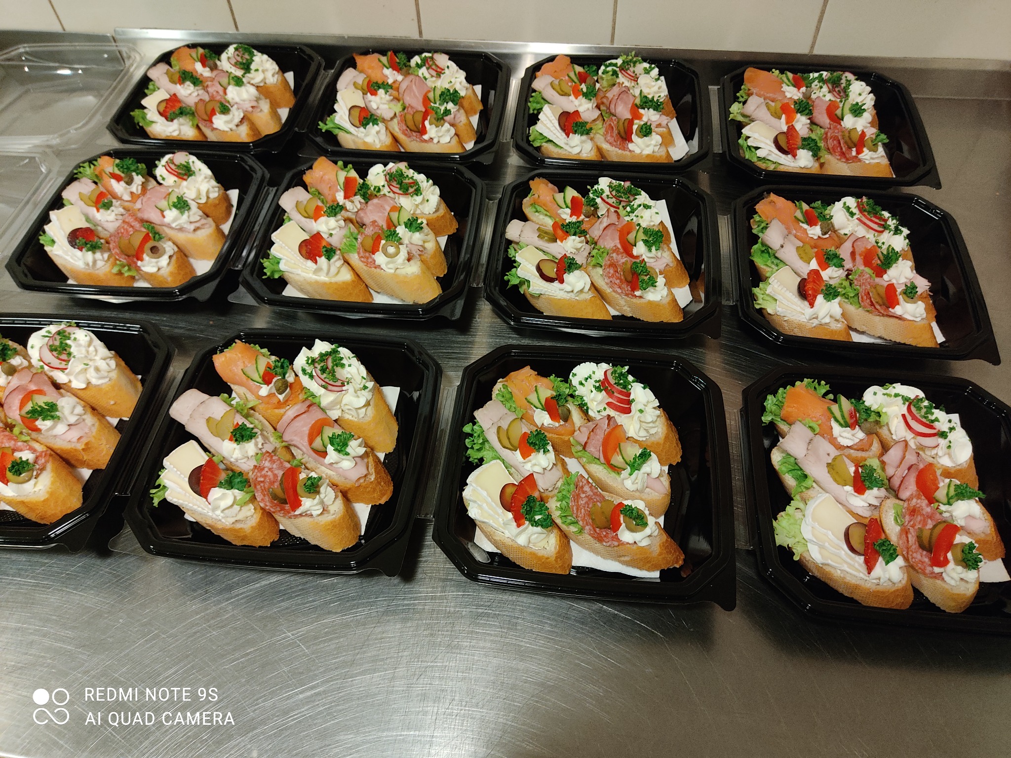 Przygotowane kanapeczki na pieczywie, udekorowane kremem, wędlinami, serem, warzywami, ułożone w czarnych, plastikowych tackach na metalowym stole.