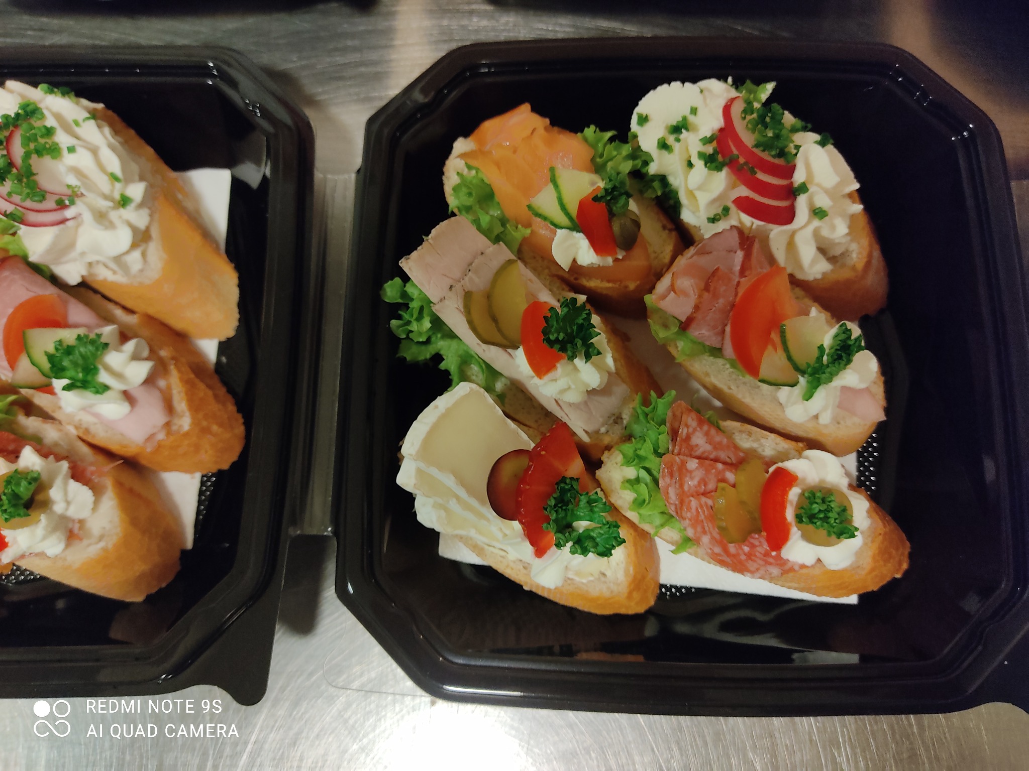 Dwie czarne tacki wypełnione różnorodnymi, apetycznie udekorowanymi kanapeczkami na pieczywie bagietkowym, z kremowym serkiem, wędlinami, łososiem, warzywami i ziołami.