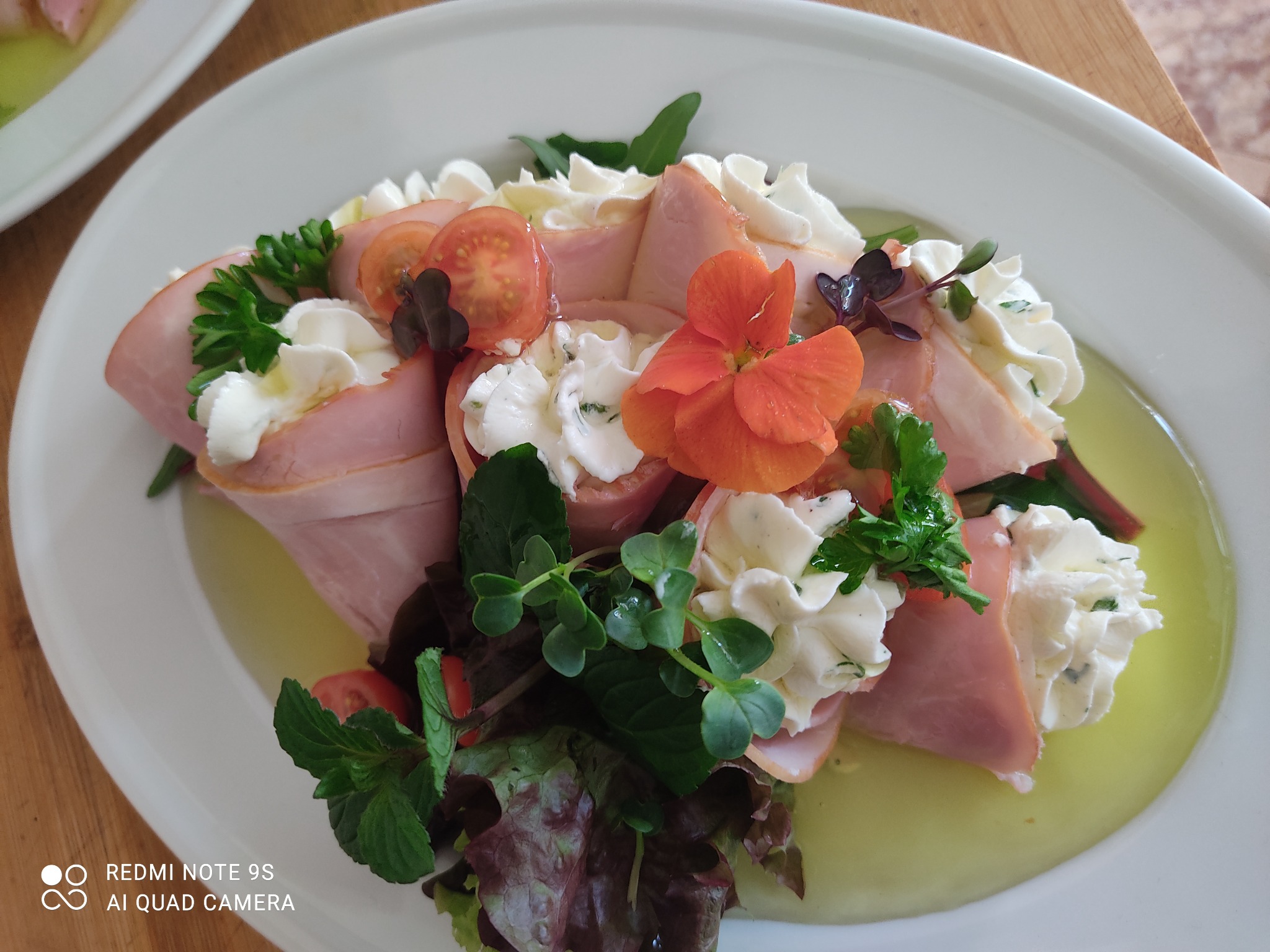 Elegancko podane roladki z szynki z kremowym serem, udekorowane pomidorkami cherry, świeżymi ziołami i jadalnym kwiatem na białym talerzu z delikatnym sosem.
