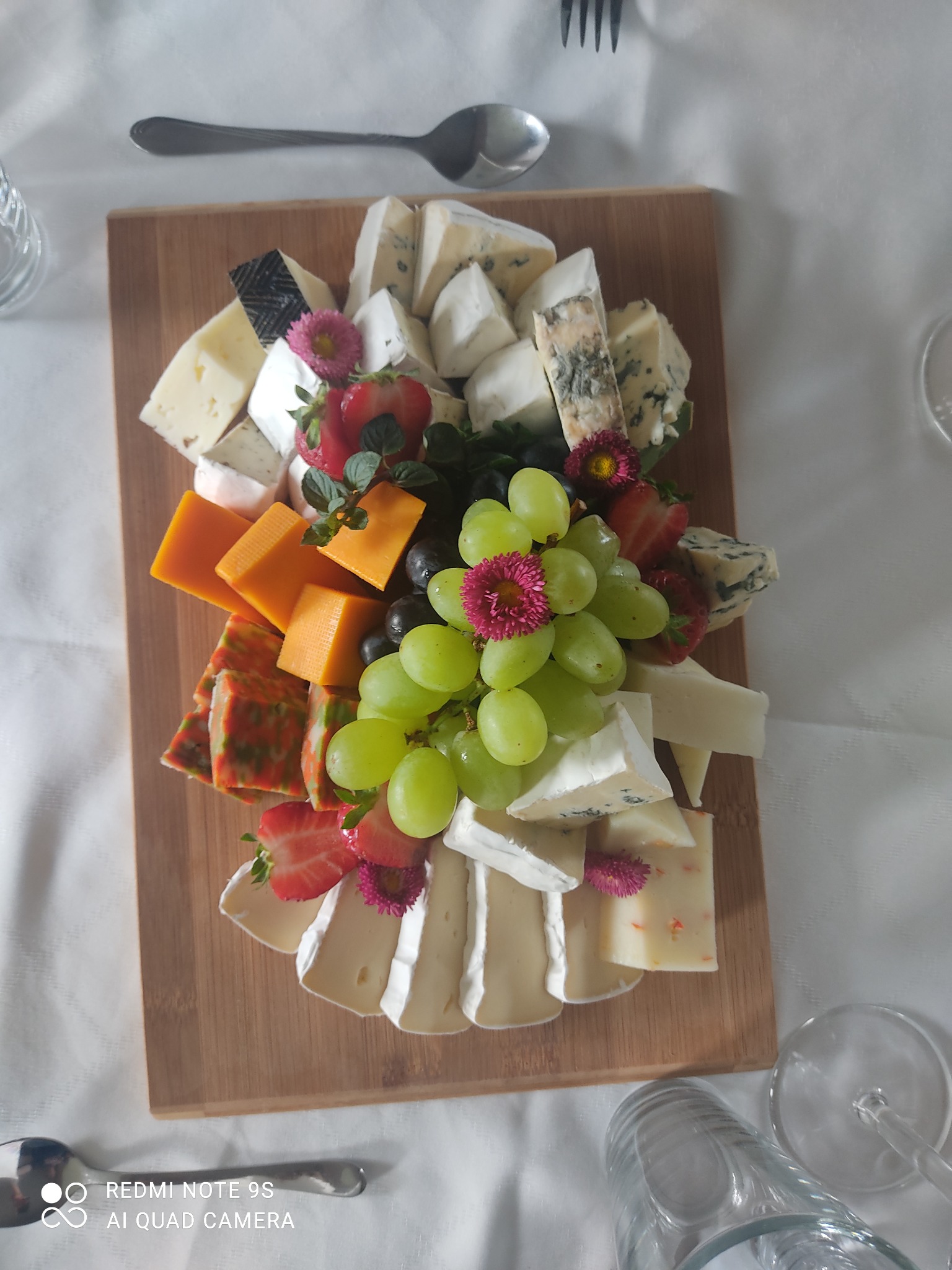 Deska serów z różnymi rodzajami sera, winogronami, truskawkami i kwiatami, ułożona na drewnianej desce na białym obrusie, widoczne sztućce i szkło.