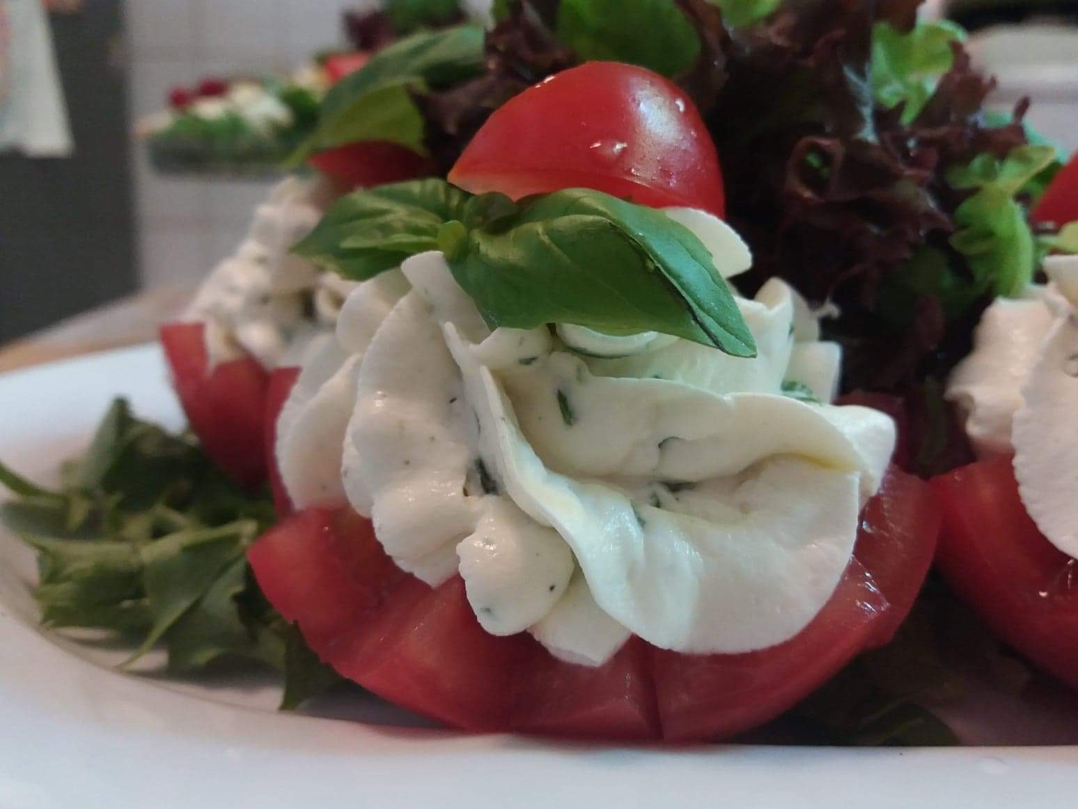 Pomidory wydrążone i wypełnione kremowym farszem ziołowym, udekorowane listkami bazylii i czerwoną cząstką pomidora, ułożone na talerzu z zieloną sałatą.