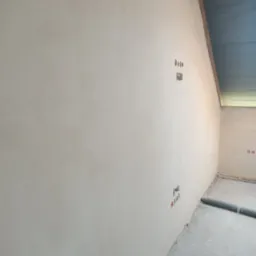 Gładka, jasna ściana po nałożeniu tynku maszynowego, z widocznymi puszkami elektrycznymi i fragmentem węża wentylacyjnego przy podłodze.