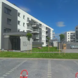 Nowoczesny budynek mieszkalny z zielonym trawnikiem, młodymi drzewkami i kamieniami ozdobnymi na pierwszym planie, widok z poziomu kostki brukowej parkingu.