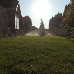 Automatyczne zraszacze nawadniające trawnik przed domem z kamienną elewacją w słoneczny dzień.