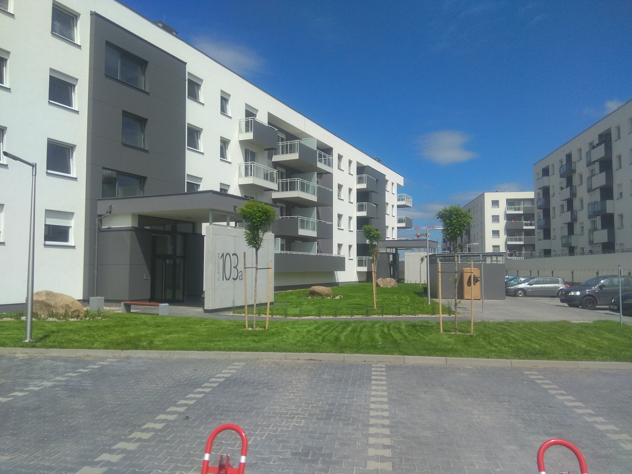 Nowoczesny budynek mieszkalny z zielonym trawnikiem, młodymi drzewkami i kamieniami ozdobnymi na pierwszym planie, widok z poziomu kostki brukowej parkingu.