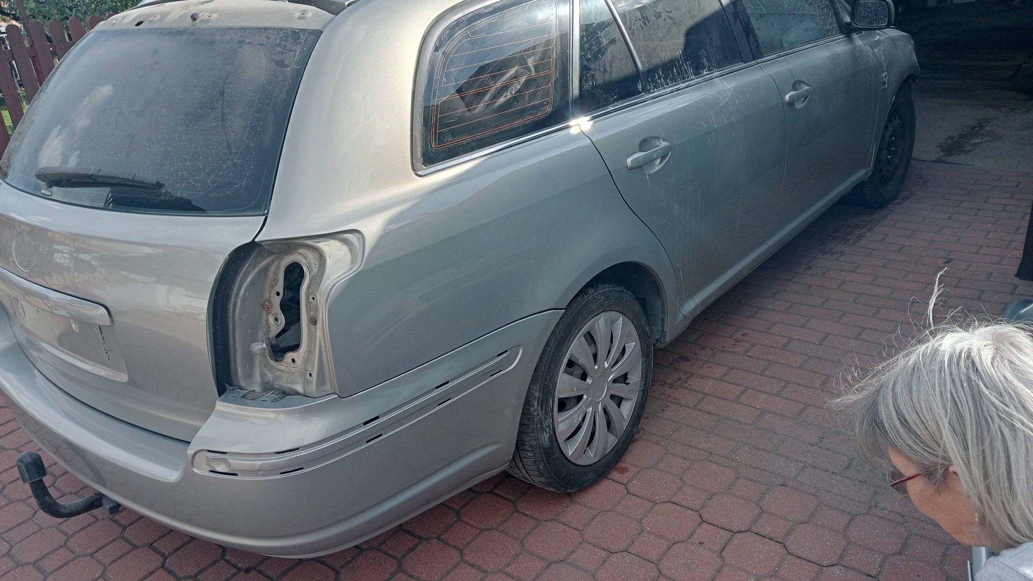 Srebrne kombi z uszkodzonym tylnym światłem, widoczne wnętrze klosza, osoba z siwymi włosami w prawym dolnym rogu kadru.