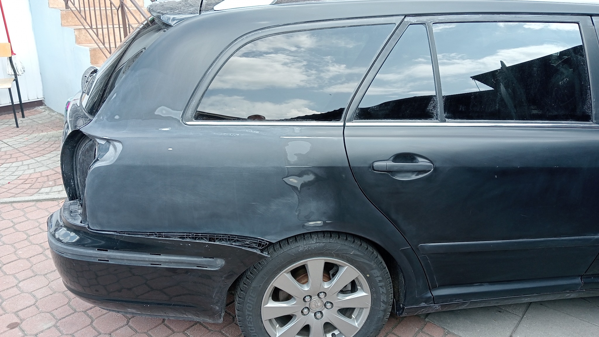 Ciemny samochód kombi po kolizji, w trakcie naprawy blacharskiej, z widocznymi śladami szpachli i uszkodzeniami tylnego zderzaka. W szybach odbija się niebo.