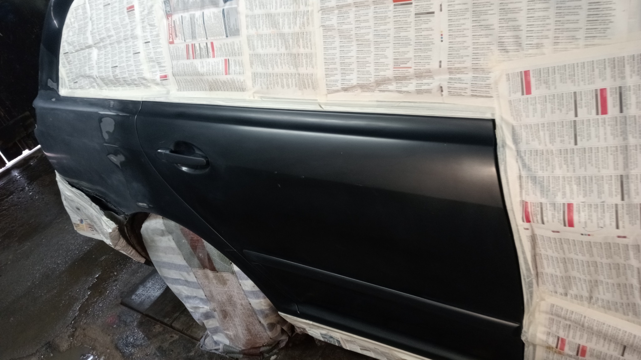 Przygotowanie do lakierowania boku samochodu, zabezpieczonego gazetami, widoczne niedoskonałości na powierzchni.