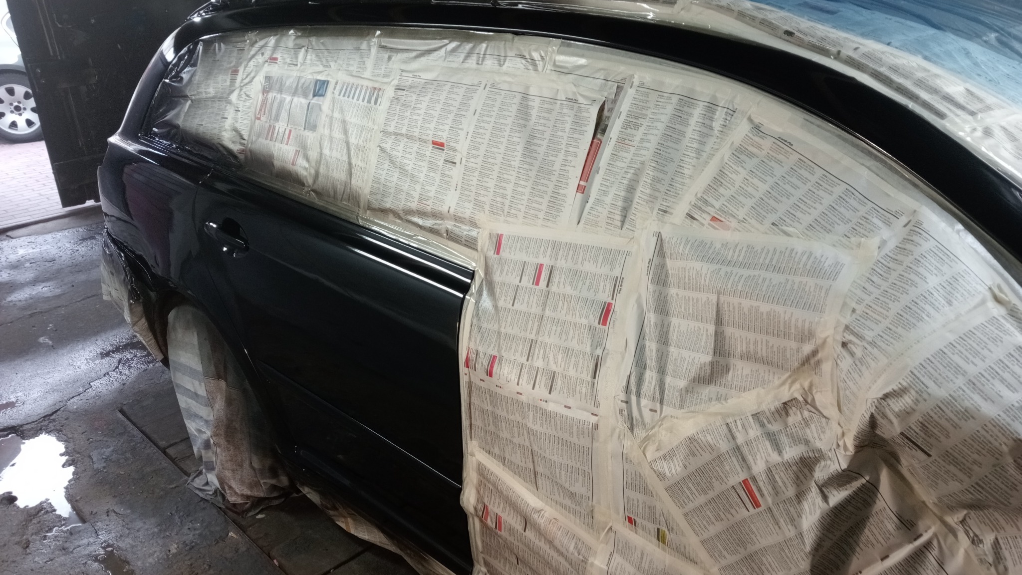 Ciemny samochód osobowy w trakcie przygotowania do lakierowania, oklejony gazetami i folią ochronną w warsztacie.