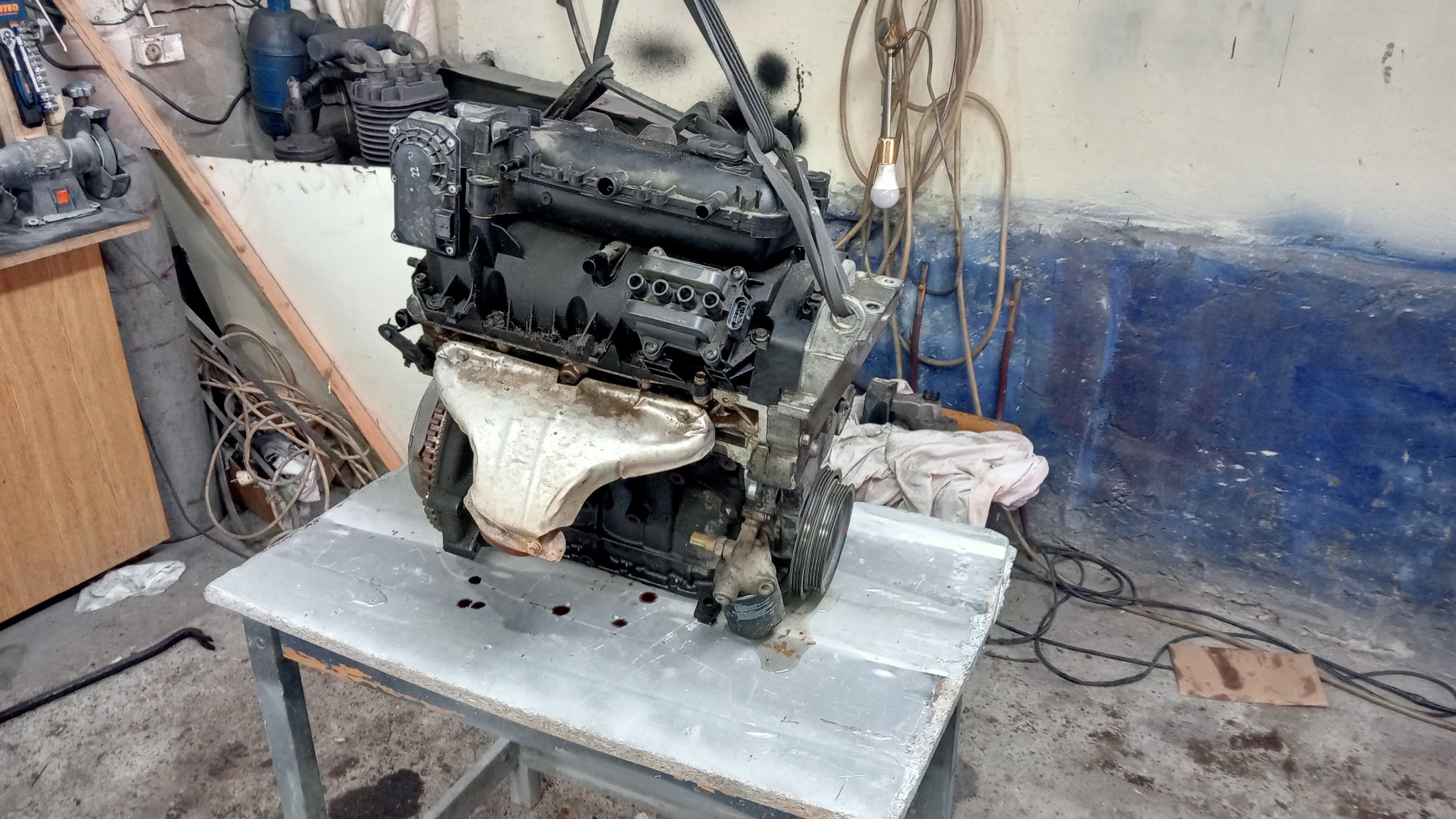 Silnik samochodowy zawieszony na pasach, umieszczony na metalowym stole w warsztacie; widoczne ślady oleju i zabrudzenia.