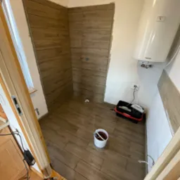 Wnętrze łazienki w Ustroniu w trakcie remontu, widoczne brązowe płytki imitujące drewno na ścianach i podłodze, biały bojler na ścianie, narzędzia w czarnym pojemniku oraz wiadro z zaprawą.