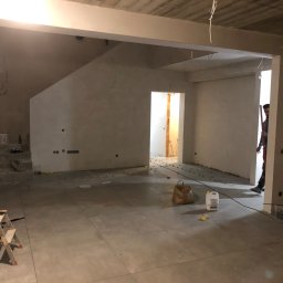 Gomola - Usługi glazurnicze - Wnętrze w trakcie prac wykończeniowych, z ułożonymi płytkami podłogowymi, schodami w tle i widocznymi narzędziami oraz materiałami budowlanymi, w tym poziomica na schodach i wiadro z zaprawą w rogu...