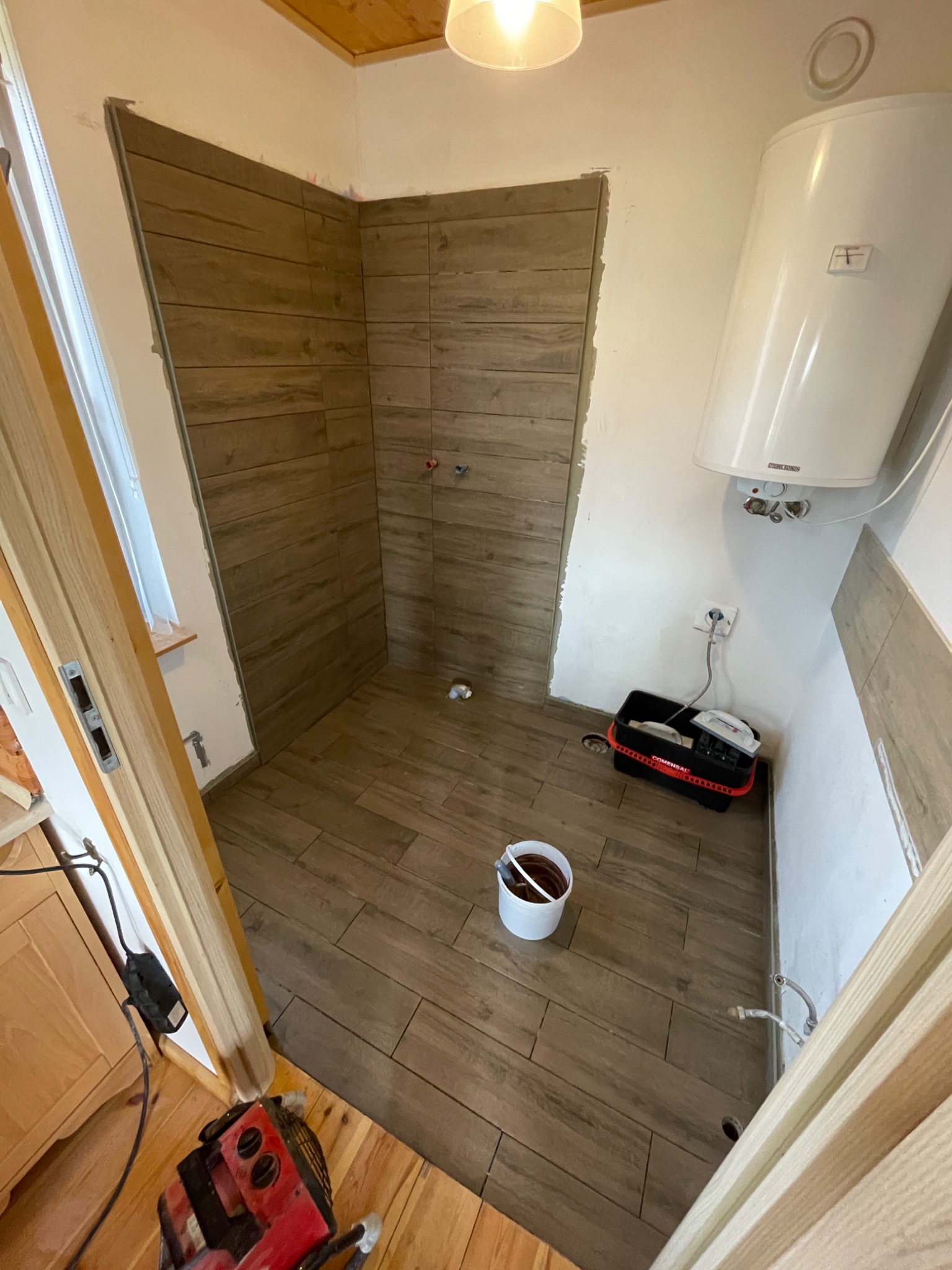 Wnętrze łazienki w Ustroniu w trakcie remontu, widoczne brązowe płytki imitujące drewno na ścianach i podłodze, biały bojler na ścianie, narzędzia w czarnym pojemniku oraz wiadro z zaprawą.